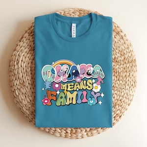 Puede incluir: Camiseta de color turquesa con la frase "Ohana Means Family" en una fuente retro colorida. El dise&ntilde;o incluye un arco&iacute;ris, flores y estrellas. La camiseta est&aacute; doblada sobre una superficie circular tejida.
