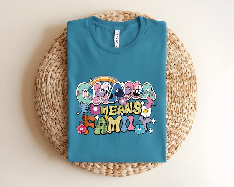 Puede incluir: Camiseta de color turquesa con la frase "Ohana Means Family" en una fuente retro colorida. El dise&ntilde;o incluye un arco&iacute;ris, flores y estrellas. La camiseta est&aacute; doblada sobre una superficie circular tejida.
