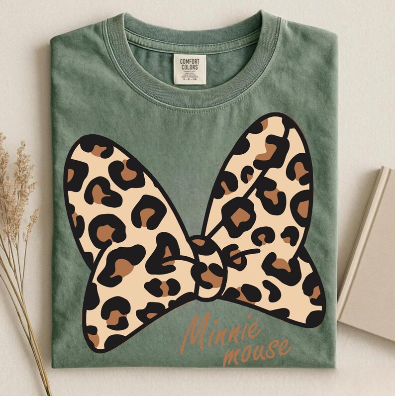 May include: A sage green t-shirt featuring a large bow design with a leopard print pattern in tan and brown. The words "Minnie mouse" are printed in a cursive font below the bow. The shirt has a crew neck and short sleeves.