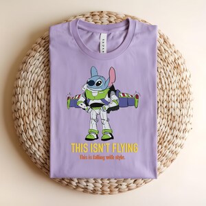 May include: A lilac-colored t-shirt featuring a cartoon character in a Buzz Lightyear costume. The graphic includes the text "THIS ISN'T FLYING" in yellow. The shirt is folded on a woven, circular mat.