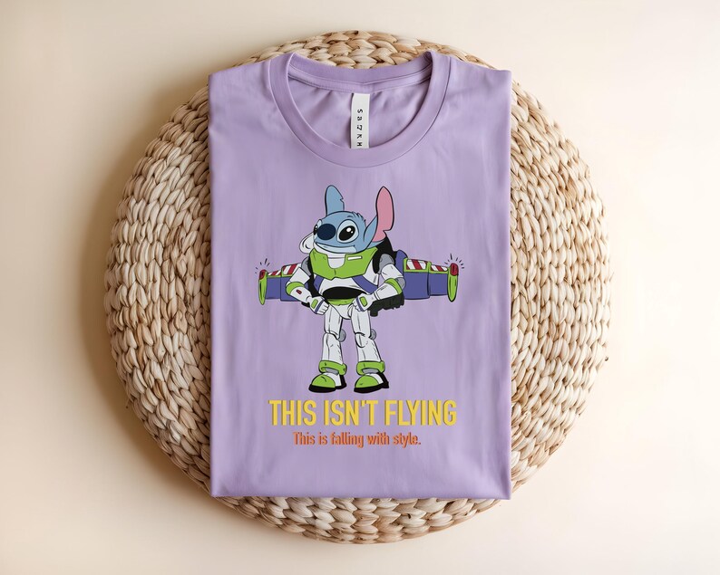 May include: A lilac-colored t-shirt featuring a cartoon character in a Buzz Lightyear costume. The graphic includes the text "THIS ISN'T FLYING" in yellow. The shirt is folded on a woven, circular mat.