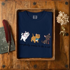 May include: Navy blue t-shirt with a cartoon cat design and the text "everybody wants to be a cat". The shirt is presented in a wooden tray. The design features three cats with bows.