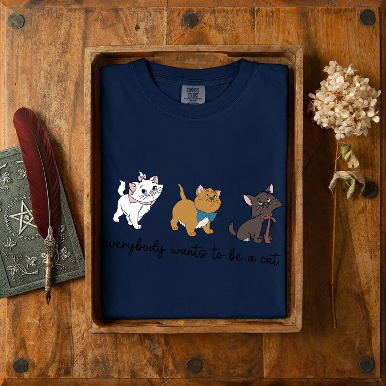May include: Navy blue t-shirt with a cartoon cat design and the text "everybody wants to be a cat". The shirt is presented in a wooden tray. The design features three cats with bows.