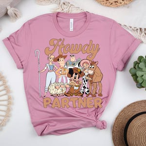 May include: A dusty rose t-shirt featuring a graphic of cartoon characters with the text "Howdy Partner" in a tan font. The design includes characters like Woody, Buzz Lightyear, and Mickey Mouse. The shirt is made of soft material.