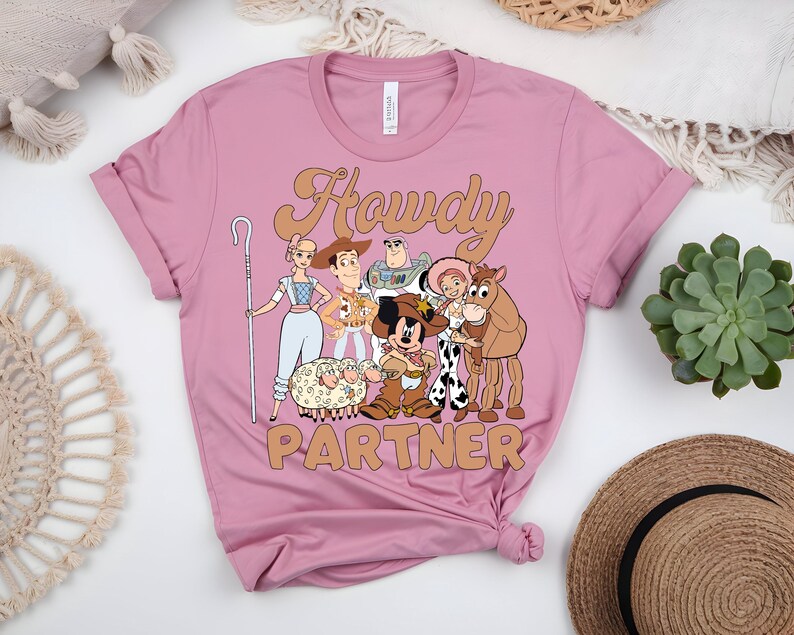 May include: A dusty rose t-shirt featuring a graphic of cartoon characters with the text "Howdy Partner" in a tan font. The design includes characters like Woody, Buzz Lightyear, and Mickey Mouse. The shirt is made of soft material.