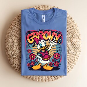 May include: A blue t-shirt featuring a cartoon of a duck with a pink bow and vest, surrounded by flowers. The word "GROOVY" is written above the duck in a colorful, retro font. The shirt is folded on a woven mat.