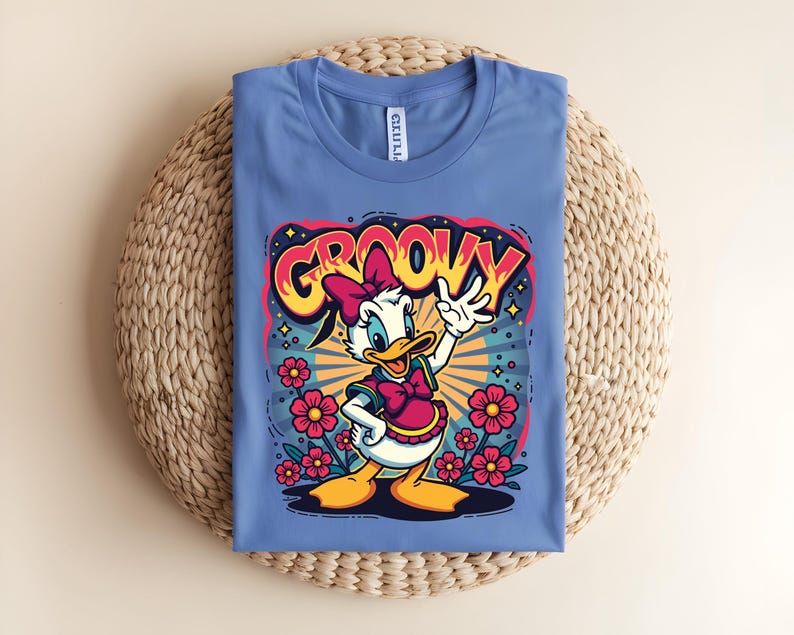 May include: A blue t-shirt featuring a cartoon of a duck with a pink bow and vest, surrounded by flowers. The word "GROOVY" is written above the duck in a colorful, retro font. The shirt is folded on a woven mat.