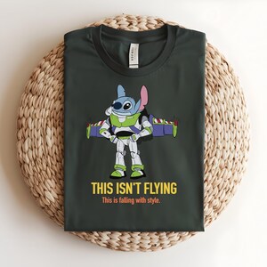 May include: Dark green t-shirt featuring a cartoon character fusion of Stitch and Buzz Lightyear. The character is wearing a Buzz Lightyear spacesuit with Stitch's features. The text "THIS ISN'T FLYING" and "This is falling with style" are printed below the character.