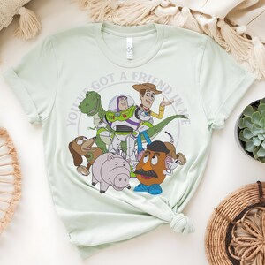 May include: Pale green t-shirt with a graphic of characters from the animated film "Toy Story." The design includes Woody, Buzz Lightyear, Rex, Mr. Potato Head, Hamm, and Slinky Dog, with the text "YOU'VE GOT A FRIEND IN ME" in a circular pattern.