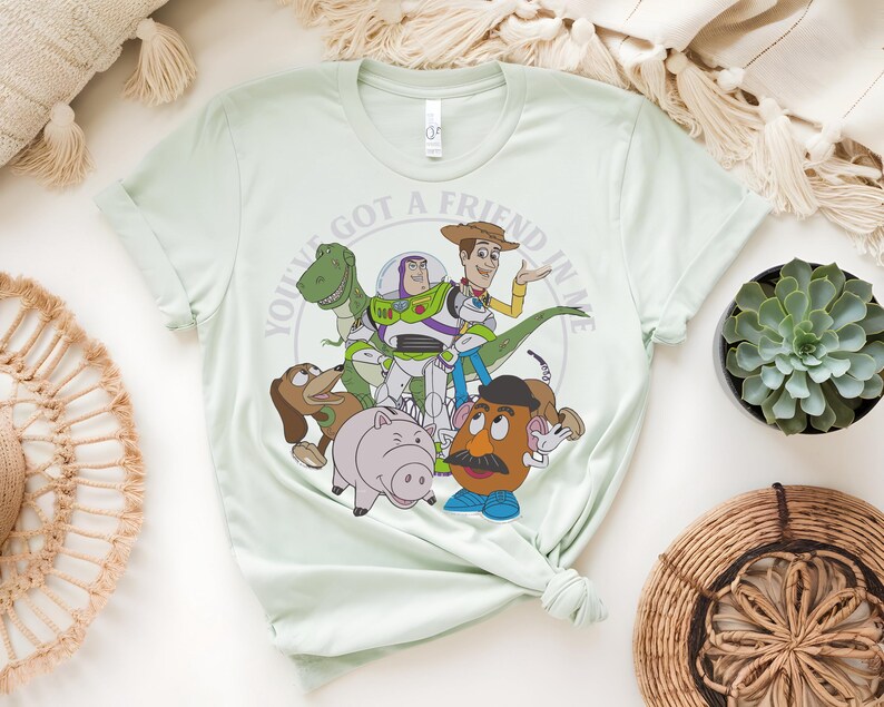 May include: Pale green t-shirt with a graphic of characters from the animated film "Toy Story." The design includes Woody, Buzz Lightyear, Rex, Mr. Potato Head, Hamm, and Slinky Dog, with the text "YOU'VE GOT A FRIEND IN ME" in a circular pattern.