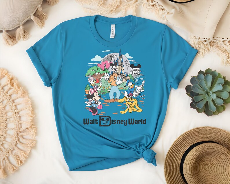 Puede incluir: Una camiseta turquesa con un gr&aacute;fico colorido de personajes de Disney, incluyendo Mickey y Minnie Mouse, Donald Duck y Goofy, frente al castillo de Cenicienta. El texto "Walt Disney World" est&aacute; impreso debajo.
