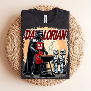 May include: Black t-shirt with a graphic of Darth Vader grilling with Stormtroopers. The design includes the text "Darthlorian" and "#1 Dad" on a red apron. The shirt is displayed on a woven surface.