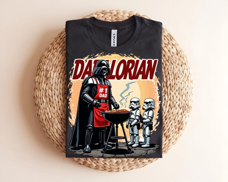 May include: Black t-shirt with a graphic of Darth Vader grilling with Stormtroopers. The design includes the text "Darthlorian" and "#1 Dad" on a red apron. The shirt is displayed on a woven surface.