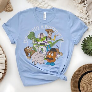 May include: Pale blue t-shirt with a graphic of Toy Story characters, including Woody, Buzz Lightyear, Rex, Slinky Dog, Hamm, and Mr. Potato Head. The text "YOU'VE GOT A FRIEND IN ME" is in a circular design around the characters.