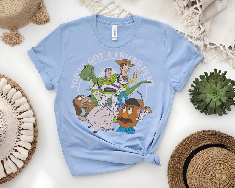 May include: Pale blue t-shirt with a graphic of Toy Story characters, including Woody, Buzz Lightyear, Rex, Slinky Dog, Hamm, and Mr. Potato Head. The text "YOU'VE GOT A FRIEND IN ME" is in a circular design around the characters.