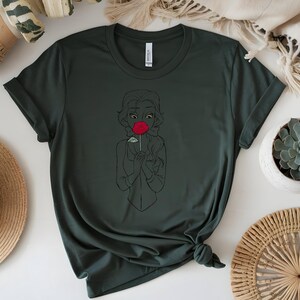 May include: Dark green t-shirt featuring a black line drawing of a person holding a red rose. The design is centered on the shirt. The shirt is knotted at the bottom. A straw hat and a succulent plant in a white pot are in the background.