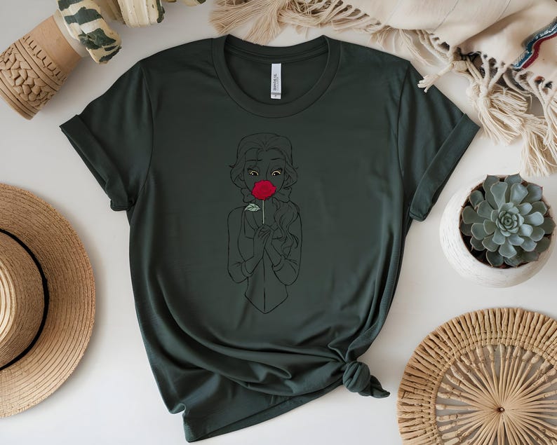 May include: Dark green t-shirt featuring a black line drawing of a person holding a red rose. The design is centered on the shirt. The shirt is knotted at the bottom. A straw hat and a succulent plant in a white pot are in the background.