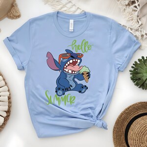 K&ouml;nnte beinhalten: Hellblaues T-Shirt mit einer Cartoonfigur, Stitch, die eine Sonnenbrille tr&auml;gt und Eis isst. Die Worte "hello summer" sind in gr&uuml;ner Schrift geschrieben. Das T-Shirt ist unten geknotet.