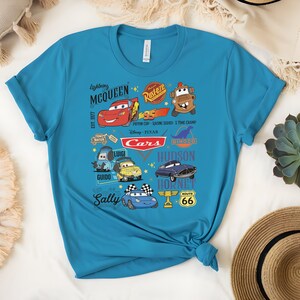 May include: A turquoise t-shirt featuring a colorful graphic of characters and logos from the Disney Pixar movie "Cars." The design includes Lightning McQueen, Mater, and other characters, along with the text "Cars."