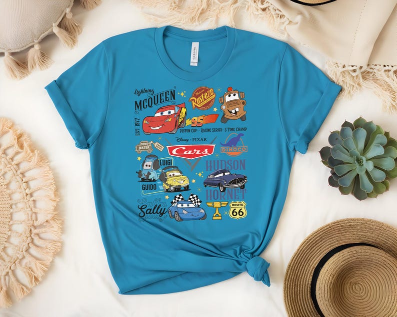 May include: A turquoise t-shirt featuring a colorful graphic of characters and logos from the Disney Pixar movie "Cars." The design includes Lightning McQueen, Mater, and other characters, along with the text "Cars."