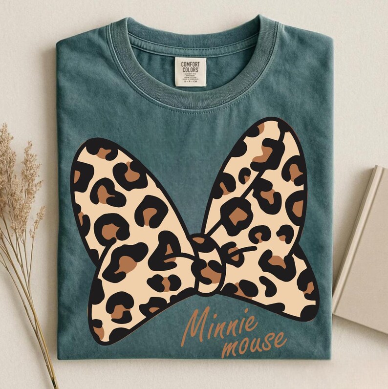 May include: A teal Comfort Colors t-shirt featuring a large leopard print bow design with the words "Minnie mouse" in a script font. The bow has tan and brown spots. The shirt is folded neatly.