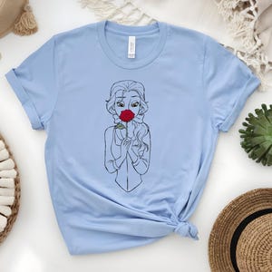 May include: Light blue t-shirt featuring a black and white line drawing of a woman holding a red rose. The illustration is centered on the shirt. The shirt is made of a soft material and has a crew neck.