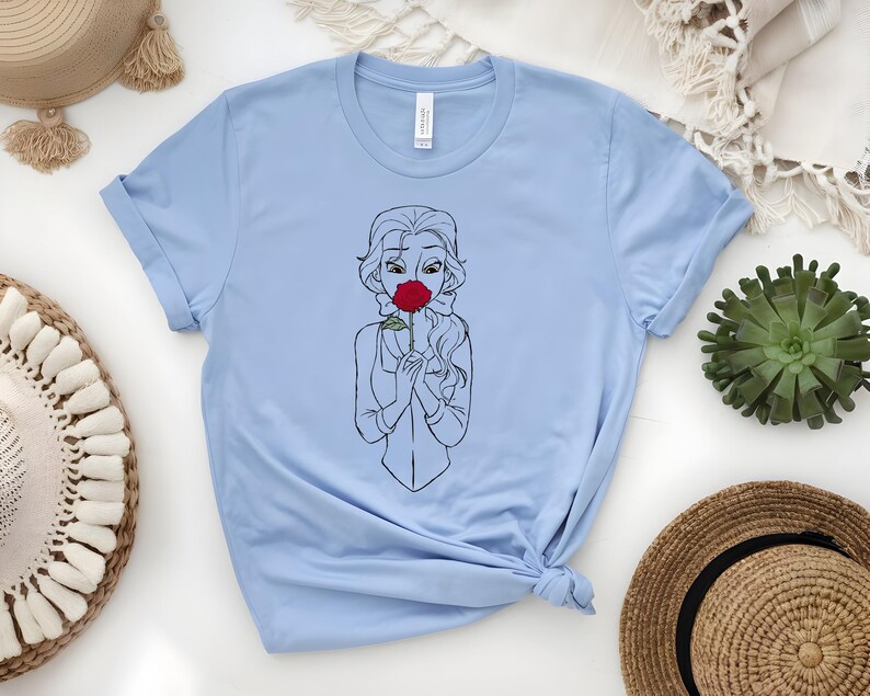 May include: Light blue t-shirt featuring a black and white line drawing of a woman holding a red rose. The illustration is centered on the shirt. The shirt is made of a soft material and has a crew neck.