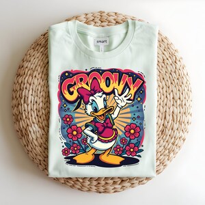 May include: A light green t-shirt featuring a cartoon graphic of a duck with a pink bow and the word "GROOVY" in a colorful design. The shirt is folded on a woven mat.