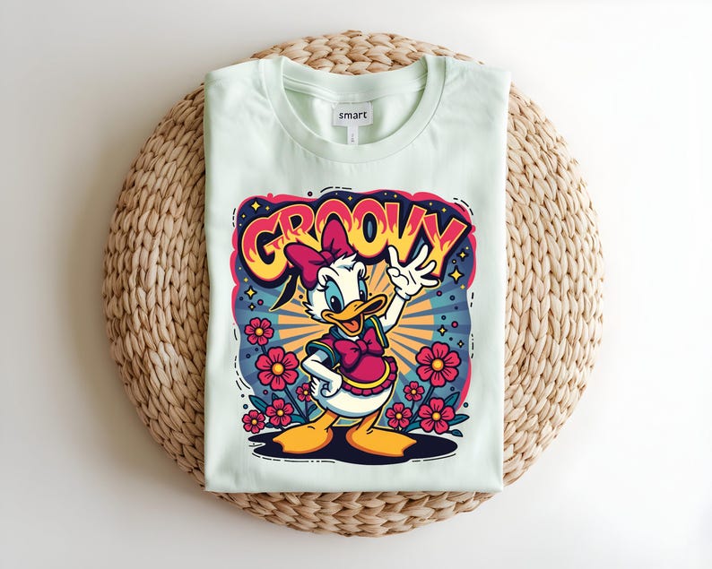 May include: A light green t-shirt featuring a cartoon graphic of a duck with a pink bow and the word "GROOVY" in a colorful design. The shirt is folded on a woven mat.