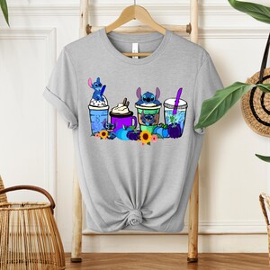 Puede incluir: Camiseta gris claro con un gr&aacute;fico colorido de personajes de Stitch con varias bebidas, incluyendo caf&eacute; y bebidas con pajitas. El dise&ntilde;o incluye girasoles y calabazas. La camiseta se muestra en un estante de madera.