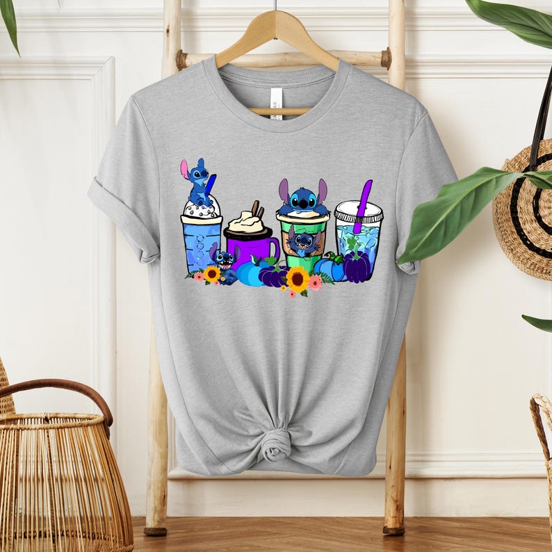Puede incluir: Camiseta gris claro con un gr&aacute;fico colorido de personajes de Stitch con varias bebidas, incluyendo caf&eacute; y bebidas con pajitas. El dise&ntilde;o incluye girasoles y calabazas. La camiseta se muestra en un estante de madera.