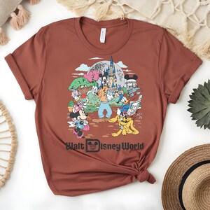 Puede incluir: Camiseta de color &oacute;xido con un gr&aacute;fico colorido de personajes de Disney, incluyendo Mickey Mouse, Minnie Mouse, Goofy, Donald Duck y Pluto, frente a un castillo. El texto "Walt Disney World" est&aacute; impreso debajo.