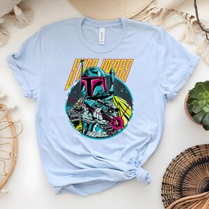 May include: Pale blue t-shirt with a vibrant graphic of a Star Wars character and the words "STAR WARS" in yellow. The design features a helmeted figure within a circular space-themed illustration.