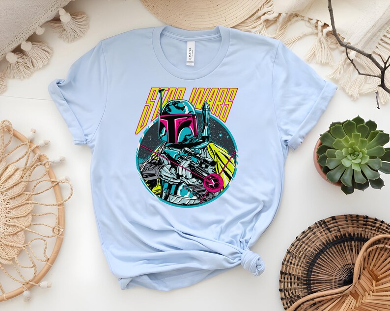 May include: Pale blue t-shirt with a vibrant graphic of a Star Wars character and the words "STAR WARS" in yellow. The design features a helmeted figure within a circular space-themed illustration.