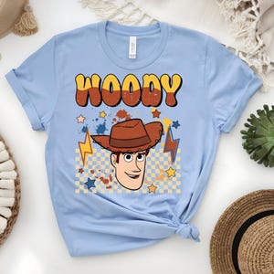 Puede incluir: Camiseta azul claro con un gr&aacute;fico de dibujos animados de Woody, con el nombre "WOODY" en una fuente retro. El dise&ntilde;o incluye un sombrero de vaquero, estrellas y rayos. La camiseta est&aacute; hecha de un material suave.