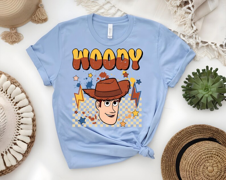 Puede incluir: Camiseta azul claro con un gr&aacute;fico de dibujos animados de Woody, con el nombre "WOODY" en una fuente retro. El dise&ntilde;o incluye un sombrero de vaquero, estrellas y rayos. La camiseta est&aacute; hecha de un material suave.