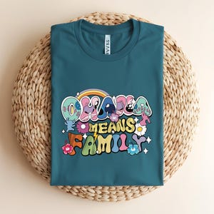 Puede incluir: Camiseta de color turquesa con la frase "Ohana Means Family" en una fuente retro colorida. El dise&ntilde;o incluye un arco&iacute;ris, flores y personajes de dibujos animados. La camiseta est&aacute; doblada sobre una estera circular tejida.