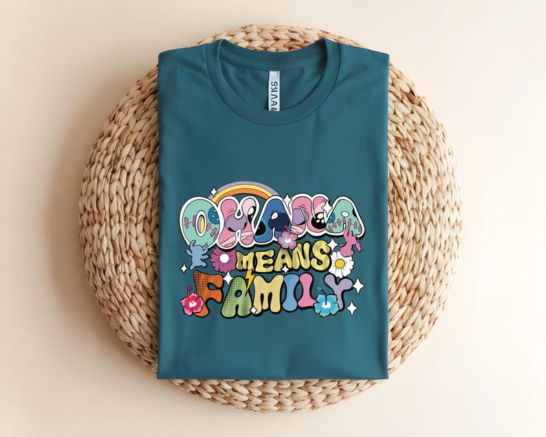 Puede incluir: Camiseta de color turquesa con la frase "Ohana Means Family" en una fuente retro colorida. El dise&ntilde;o incluye un arco&iacute;ris, flores y personajes de dibujos animados. La camiseta est&aacute; doblada sobre una estera circular tejida.