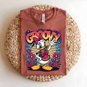 May include: A terracotta-colored t-shirt featuring a cartoon Daisy Duck design. The graphic includes the word "GROOVY" in a retro font, Daisy Duck waving, and pink flowers. The shirt is laid flat on a woven, circular surface.