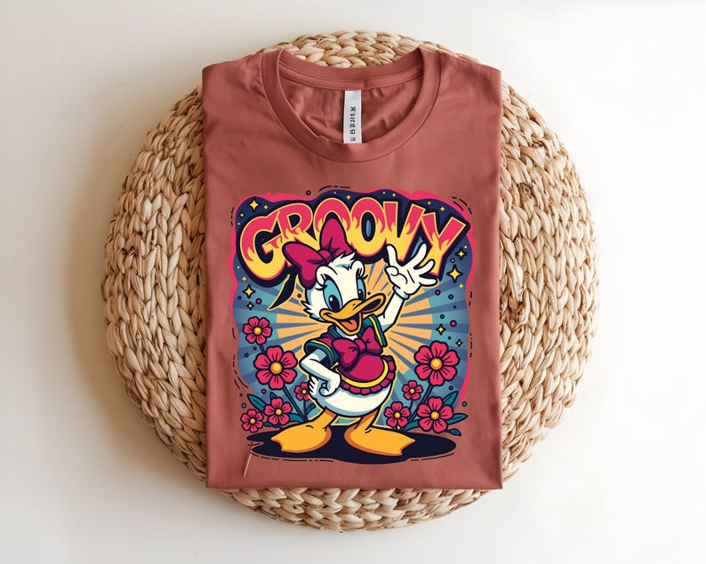 May include: A terracotta-colored t-shirt featuring a cartoon Daisy Duck design. The graphic includes the word "GROOVY" in a retro font, Daisy Duck waving, and pink flowers. The shirt is laid flat on a woven, circular surface.
