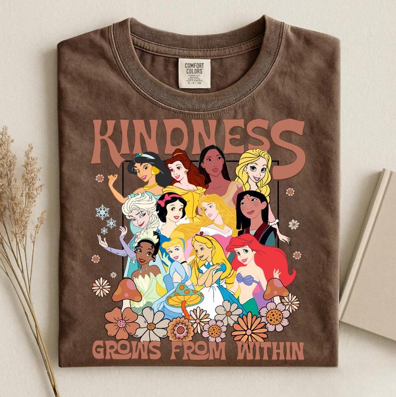 May include: Brown t-shirt featuring a graphic of various cartoon princesses with the word "KINDNESS" above them. The phrase "GROWS FROM WITHIN" is printed below the princesses, along with floral and mushroom designs.