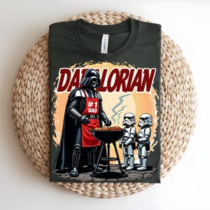 May include: Dark green t-shirt with a cartoon illustration of Darth Vader grilling. The design includes the text "Darthlorian" and "#1 Dad". Two Stormtroopers are also shown. The t-shirt is displayed on a woven surface.