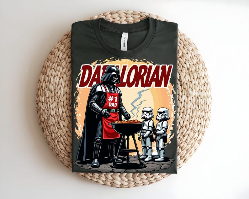 May include: Dark green t-shirt with a cartoon illustration of Darth Vader grilling. The design includes the text "Darthlorian" and "#1 Dad". Two Stormtroopers are also shown. The t-shirt is displayed on a woven surface.