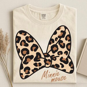 May include: Cream-colored t-shirt featuring a large bow design with a leopard print pattern in black, brown, and tan. The words "Minnie mouse" are printed in a brown script font below the bow.