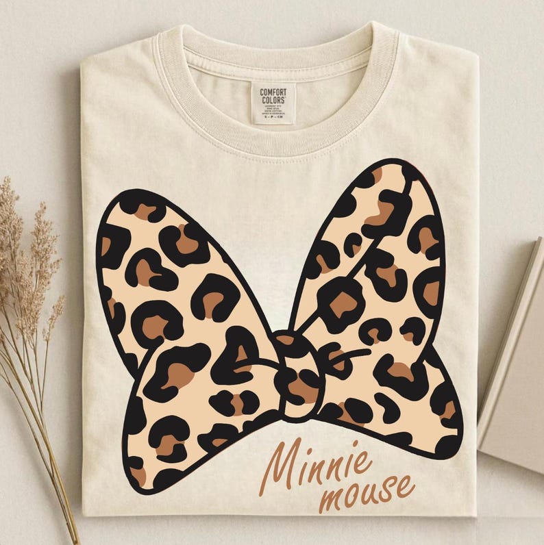 May include: Cream-colored t-shirt featuring a large bow design with a leopard print pattern in black, brown, and tan. The words "Minnie mouse" are printed in a brown script font below the bow.