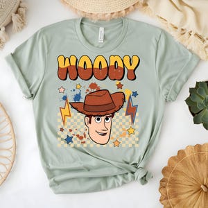 Puede incluir: Camiseta verde claro con la palabra "WOODY" en letras burbuja naranjas y marrones. Debajo, una imagen de dibujos animados de Woody con un sombrero de vaquero marr&oacute;n. El dise&ntilde;o incluye estrellas, rayos y un fondo a cuadros.
