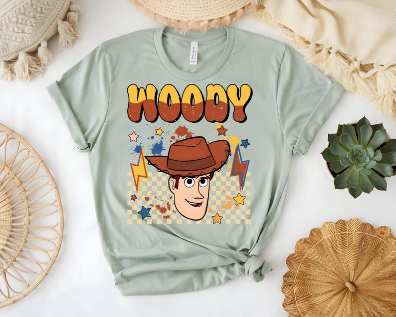Puede incluir: Camiseta verde claro con la palabra "WOODY" en letras burbuja naranjas y marrones. Debajo, una imagen de dibujos animados de Woody con un sombrero de vaquero marr&oacute;n. El dise&ntilde;o incluye estrellas, rayos y un fondo a cuadros.