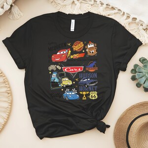 May include: Black t-shirt featuring a colorful graphic of characters and logos from the animated movie "Cars." The design includes Lightning McQueen, Mater, and the Cars logo, along with other characters and Route 66 signage.