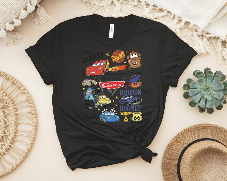 May include: Black t-shirt featuring a colorful graphic of characters and logos from the animated movie "Cars." The design includes Lightning McQueen, Mater, and the Cars logo, along with other characters and Route 66 signage.