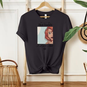 May include: Black t-shirt featuring a graphic of a red-haired character with the text "But Daddy I Love Him." The graphic is in a rectangular frame with a light blue background. The shirt is hanging on a wooden hanger.